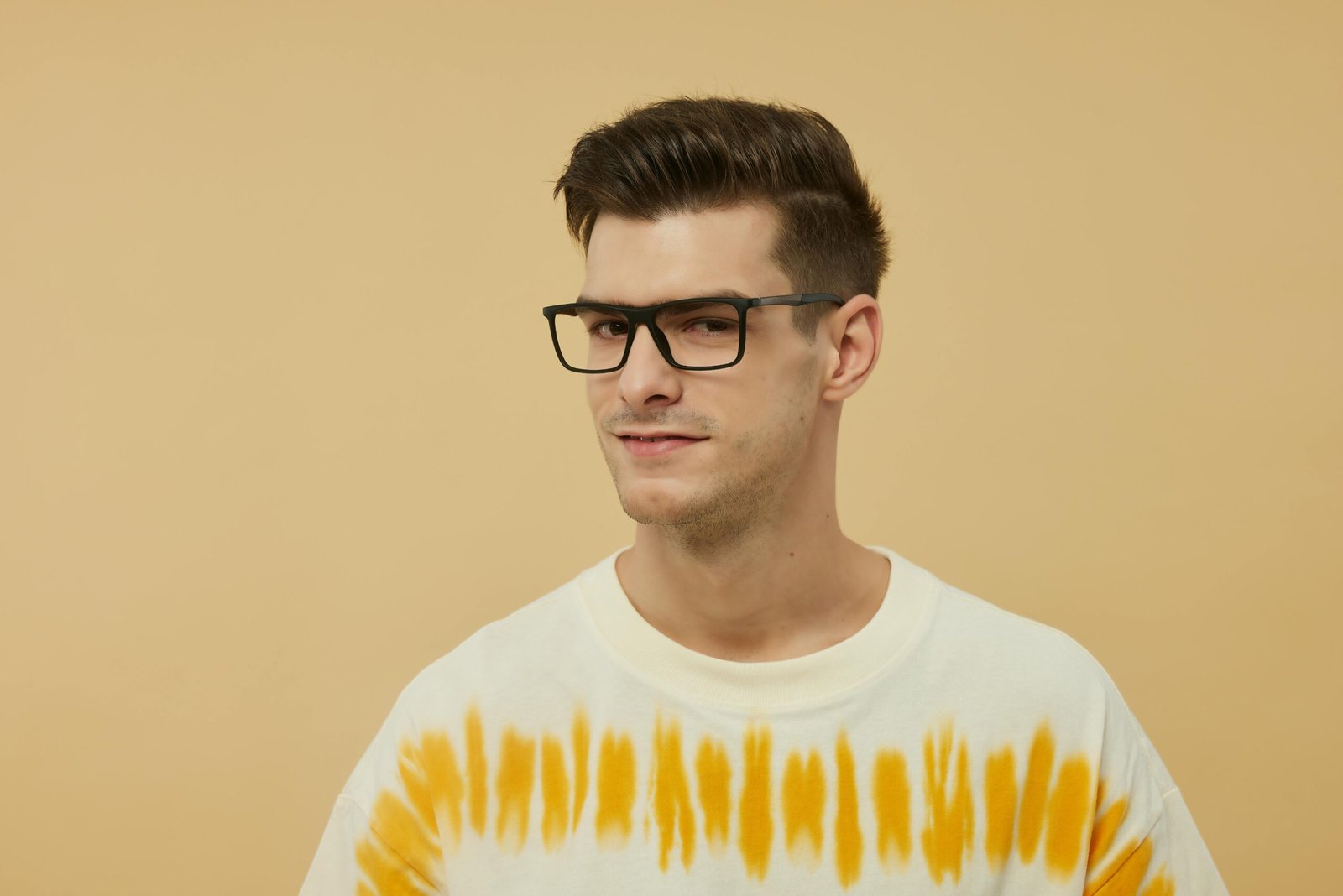 Portrait of a confident young man wearing trendy glasses and a tie-dye shirt against a beige backdrop.