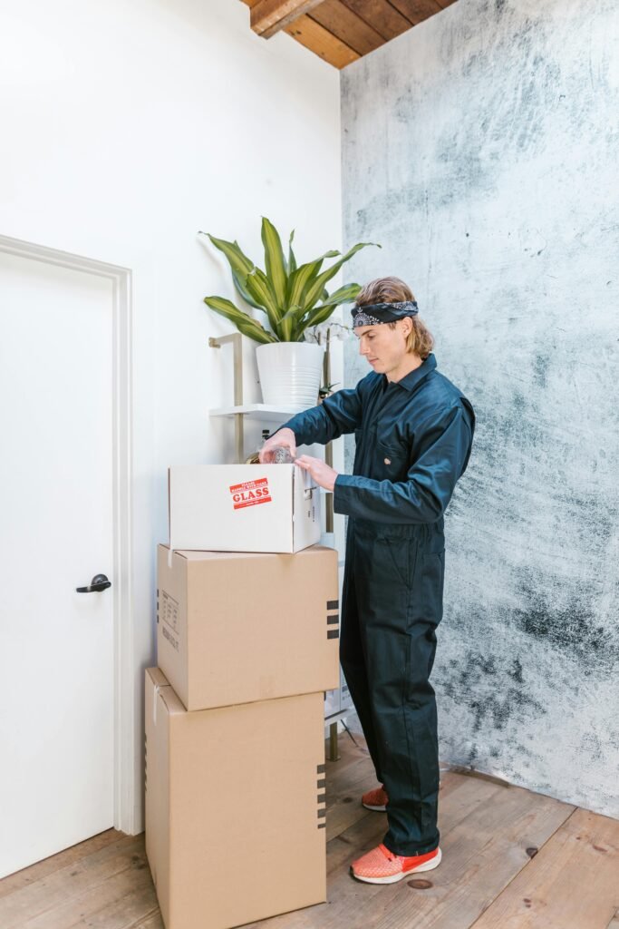 A man in coveralls stacking cardboard boxes next to a potted plant indoors.